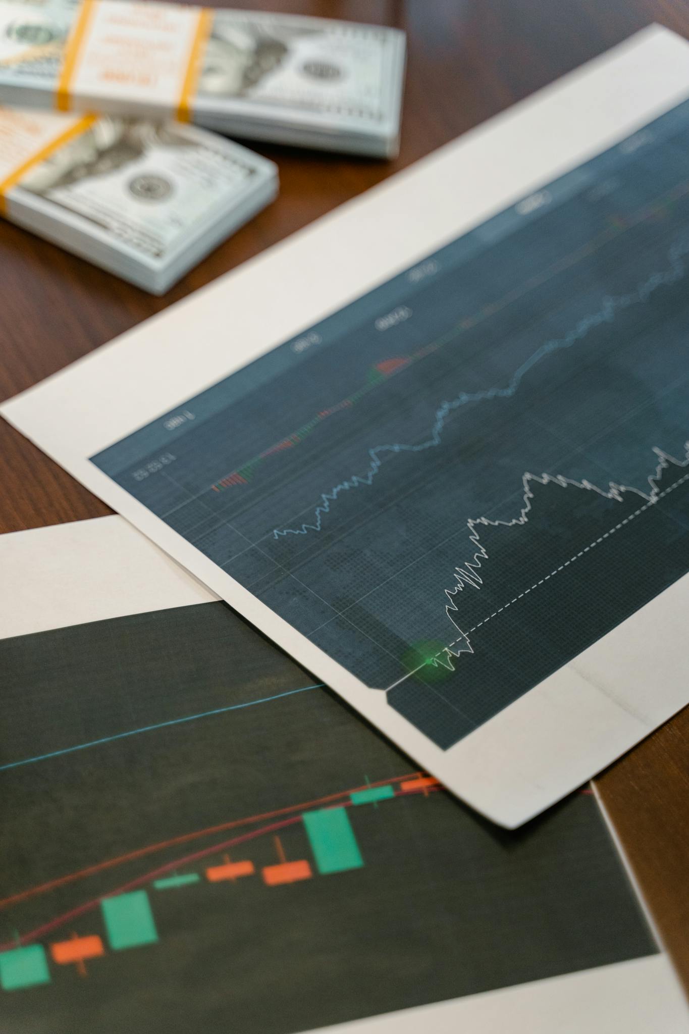 Papers with financial graphs beside stacks of US dollar bills representing market analytics.